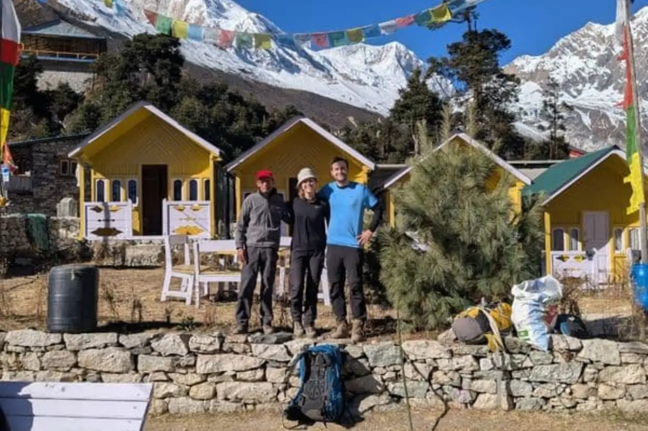 Experience Couples Romantic Nepal Trekking Annapurna A couple with a local guide standing outside yellow mountain lodges against snow peaks during a Nepal Trekking Annapurna trip.